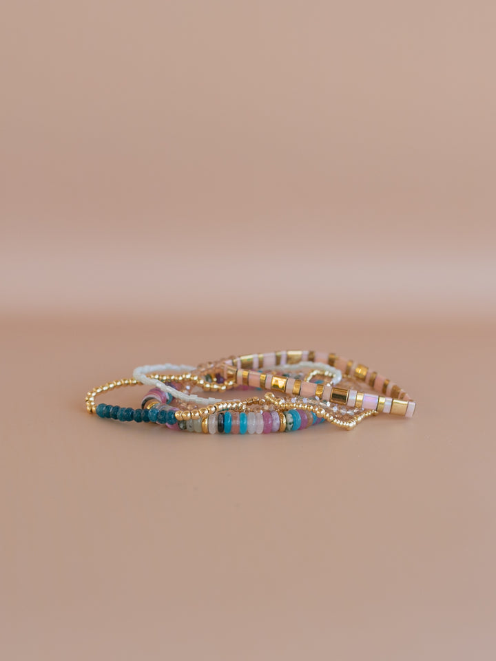 The Sapphire Bracelet stack includes gemstones, gold accents, and sparkling glass beads with a touch of pink, blue, and jewel tones. Add a pop of color to your holiday!